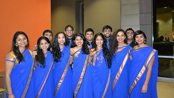 A group of women wearing blue saris with gold trim stand in front of men wearing black collared shirts and blue ties.