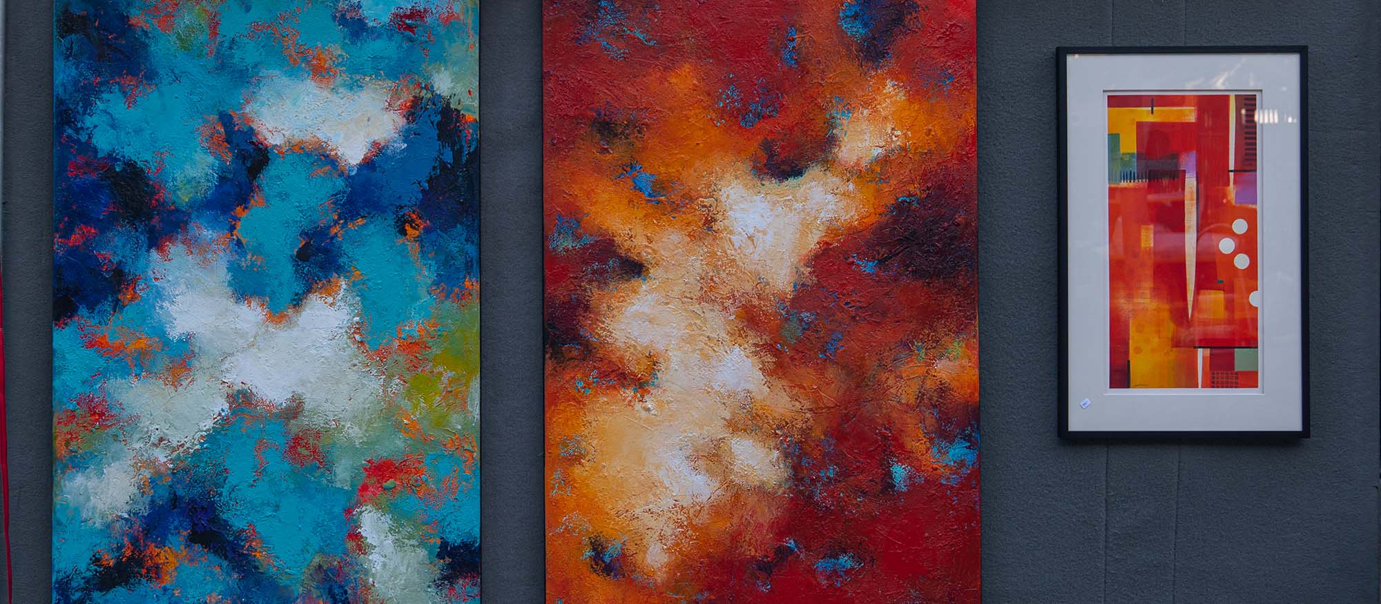 A picture of one of the booths at the art festival, with two large canvases of abstract art (one blue and one orange/red) along with a smaller framed piece of abstract art.