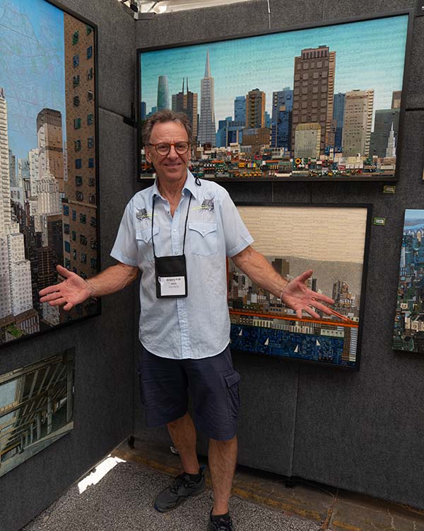 Artist Gregory Arth posing with his hands out in front of his art, which are large cityscapes.
