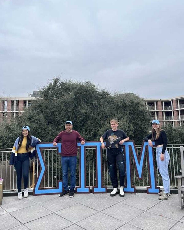 Four students stand with large Greek letters that represent Martel College - Sigma Pi Mu.