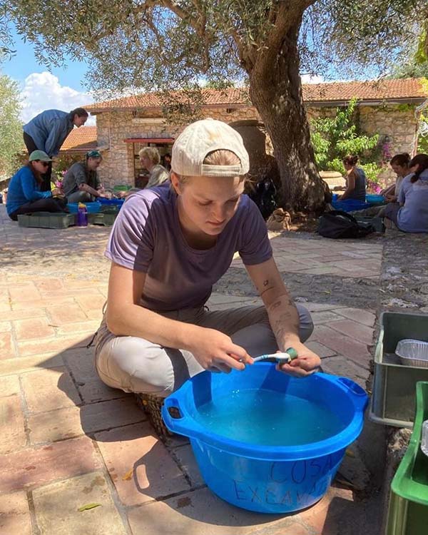 Drew is sitting behind a bucket filled with water while cleaning pieces from the archeological dig in Cosa, Italy.