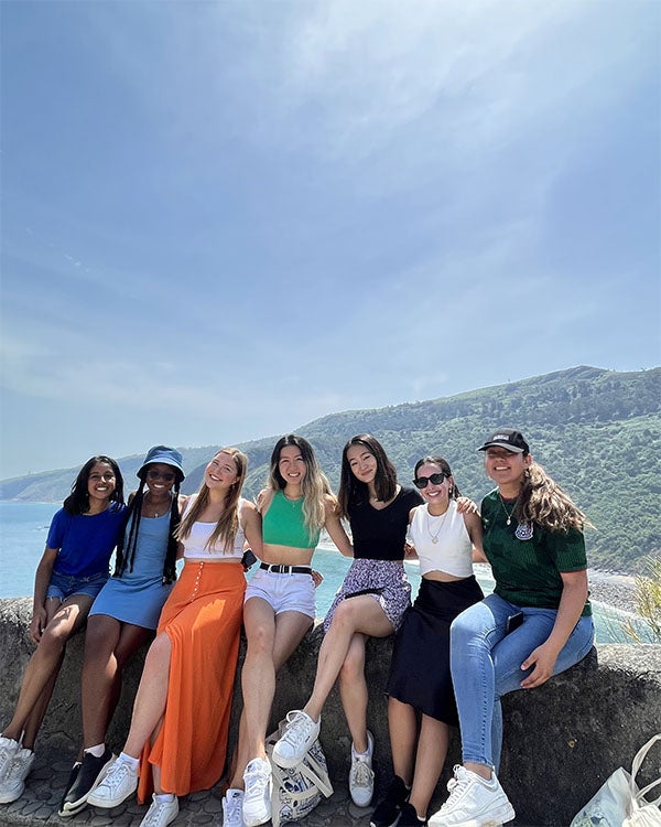 A group of seven Rice students smile for a picture while sitting on a ledge in front of a view of green-covered mountains in San Juan.