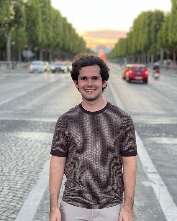 A picture of Zach Zelman, a Rice student, standing in front of a large street with tall, green trees flanking each side and a slight sunset tinge in the sky.