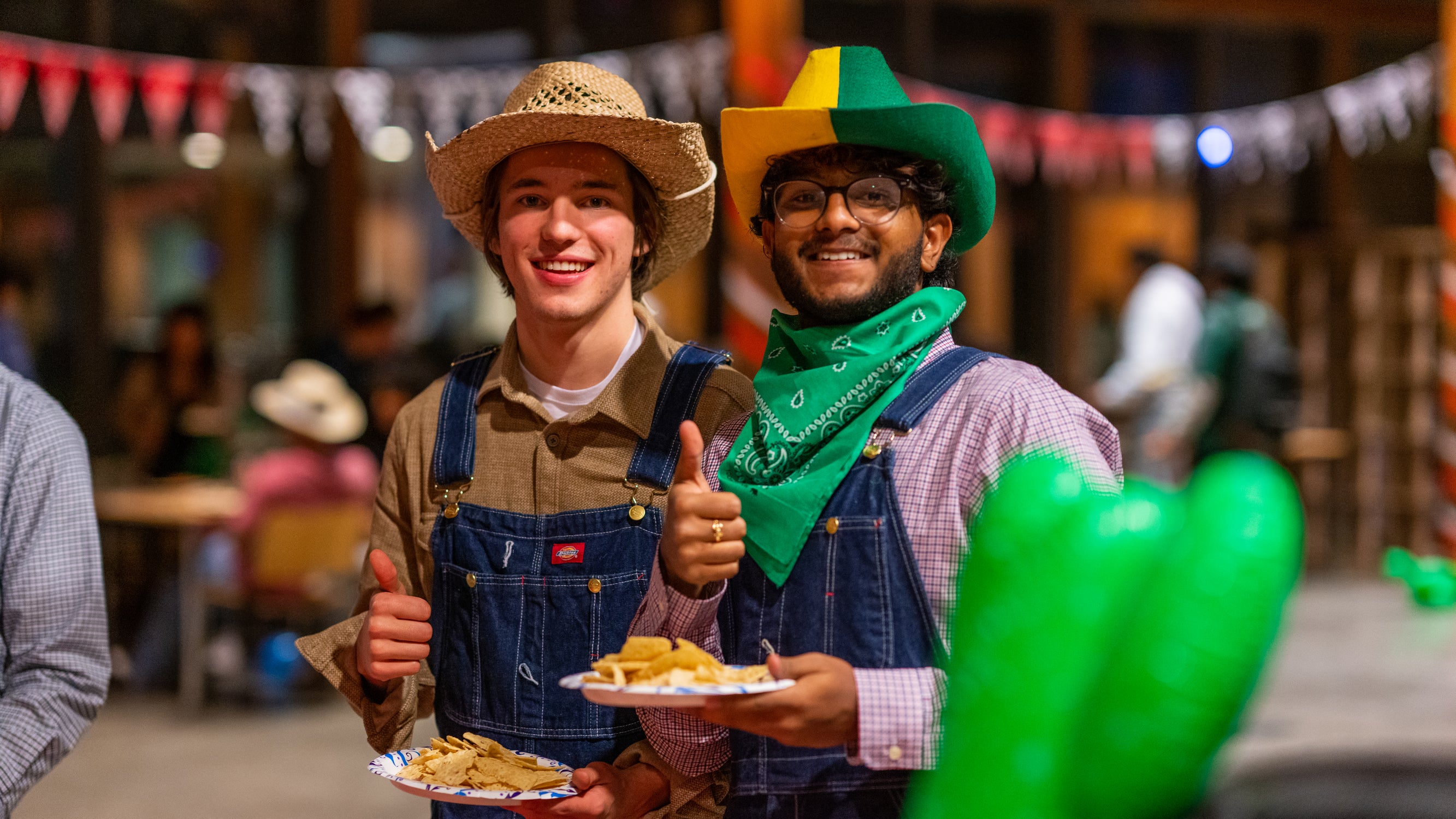 Two male students wearing cowboy hats and denim overalls holding plates of food and a thumbs up, smiling at the camera.