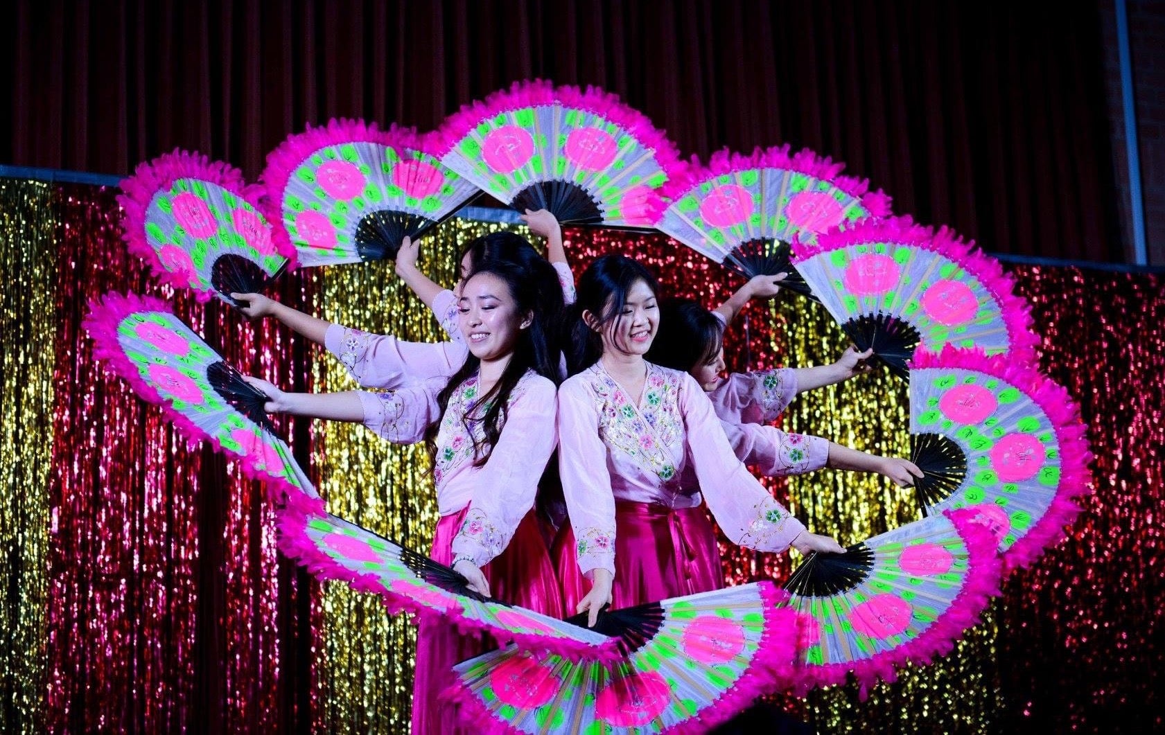 Five female dancers with bright neon pink and green fans forming a circle with the fans.