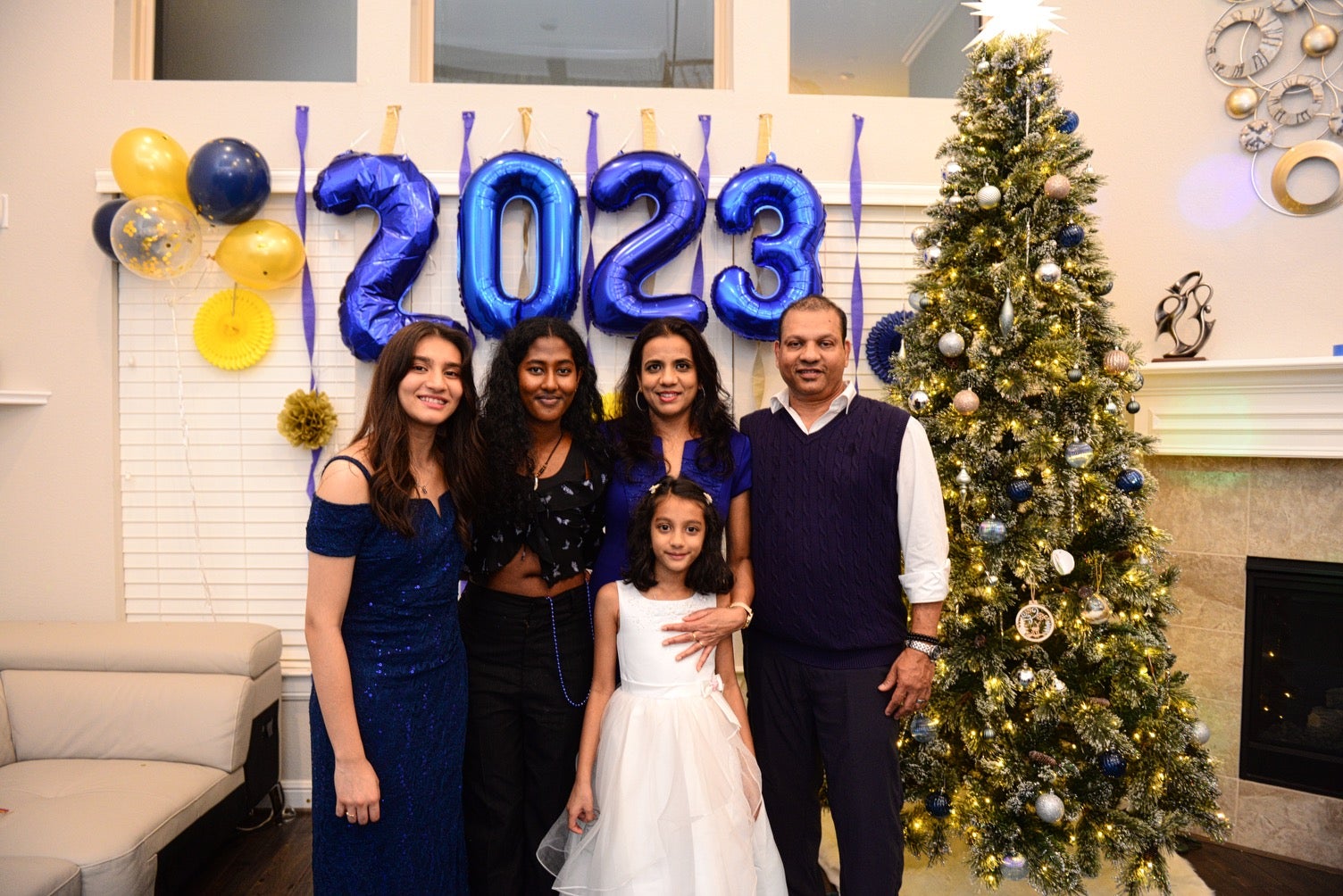 Four adults standing in a row and a child standing in front of them, smiling, with blue balloons that say "2023" and a Christmas tree behind them.