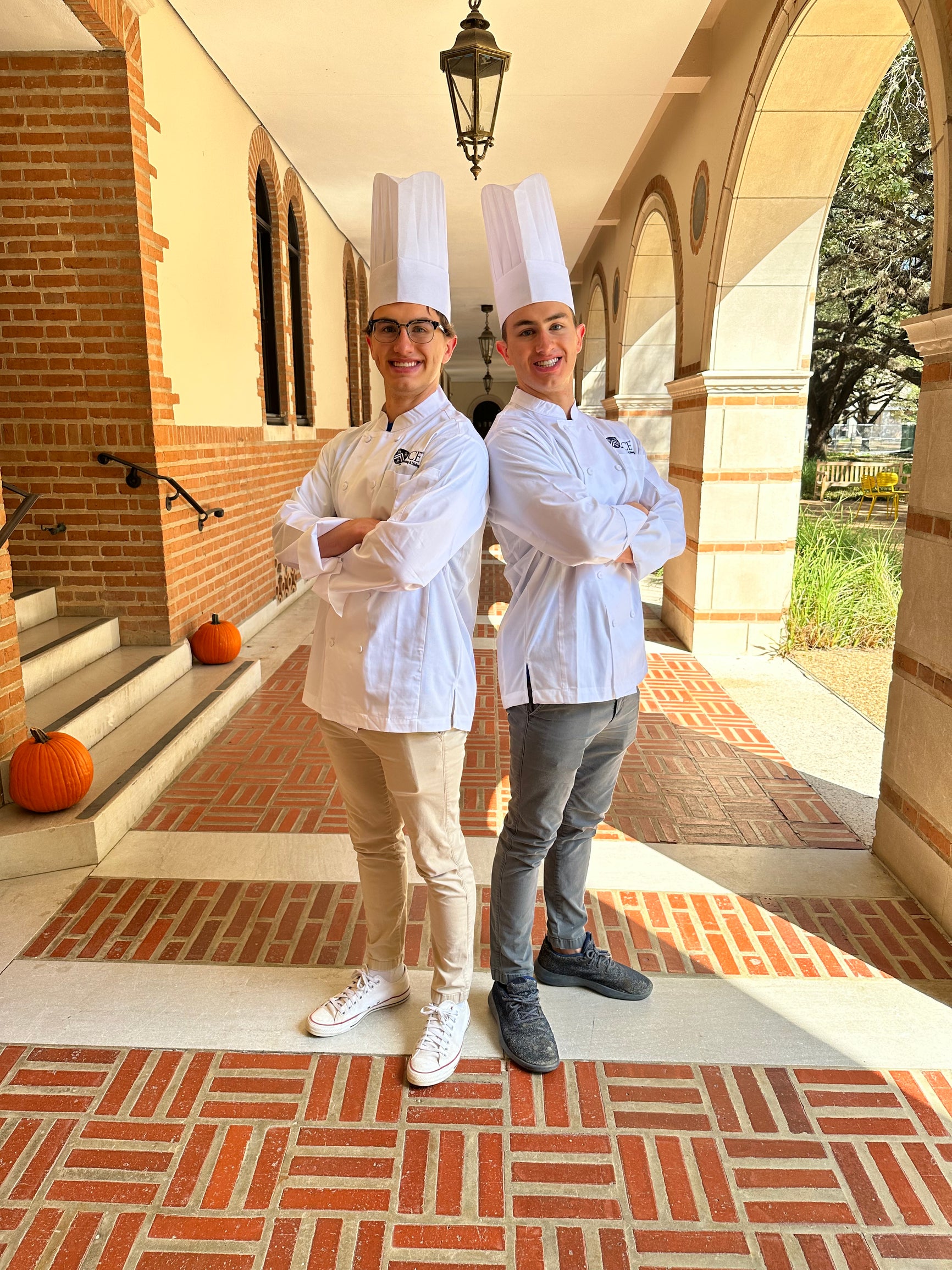 Two male students wearing a chef coat and chef hat with arms crossed in an outdoor hallway.
