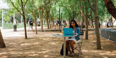 A girl sits at a table outside on Rice's campus with her laptop open in front of her.
