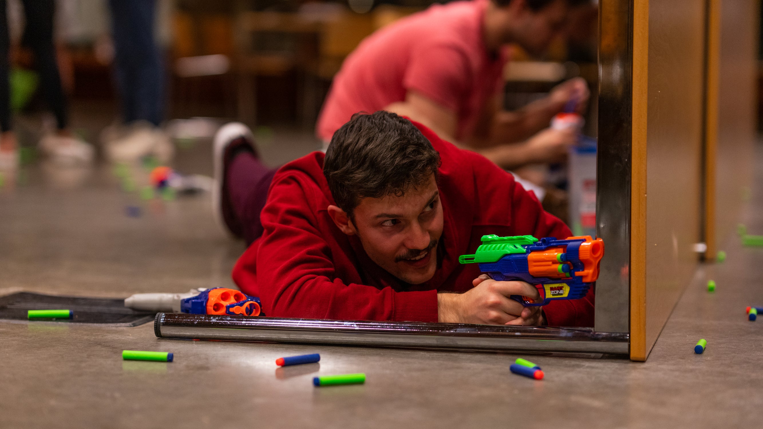 A male student laying on the floor on his stomach, hiding behind a table flipped to its side and aiming his nerf weapon.