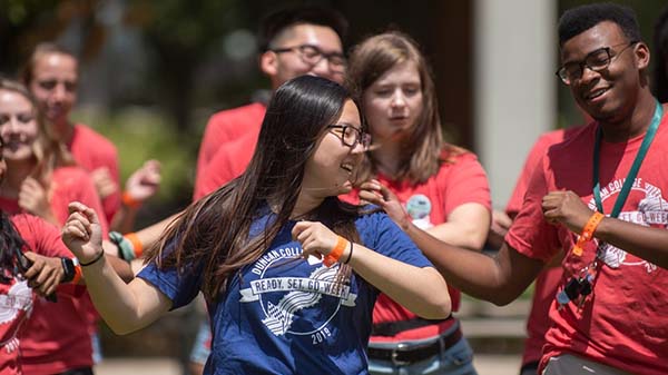 Sarah stands in the center of the frame, doing a dance move with her arm, while the students surrounding her copy her move.