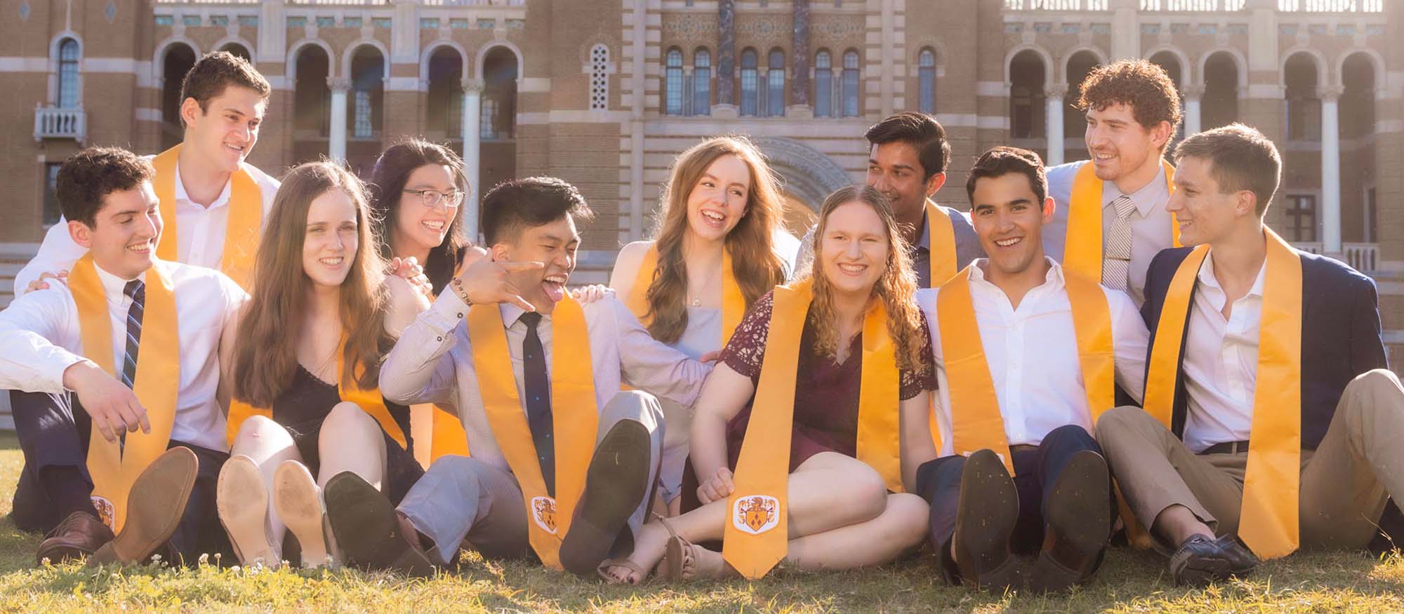 A group of students, with Pamela in the center, sit on the lawn in the academic quad wearing yellow stoles that represent Brown College.