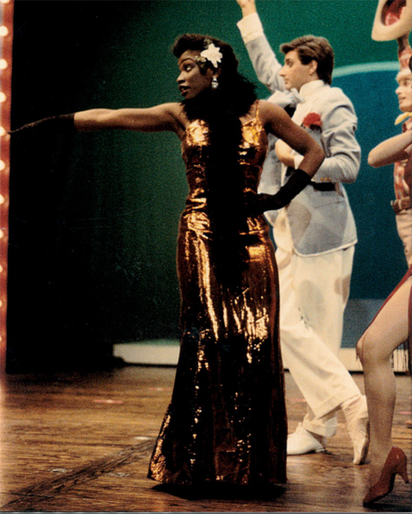 Tamara stands in the center of the frame with her arm outstretched as she performs as a character in a play.