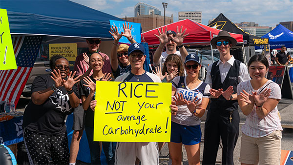 A group of students, staff and alumni pose for a picture outside of the stadium at the tailgate. The person in center is holding a sign that says “Rice: not your average carbohydrate.”