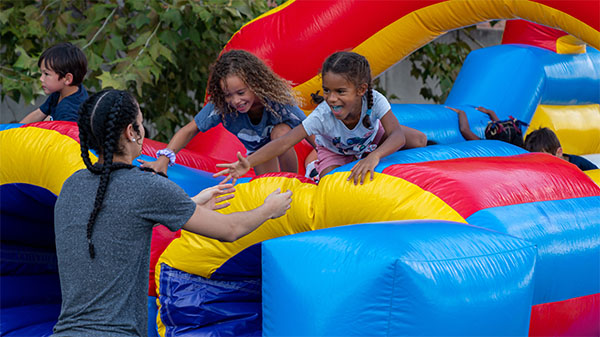 An adult stands at the end of an inflatable slide reaching towards the two children on the slide, getting ready to climb off.