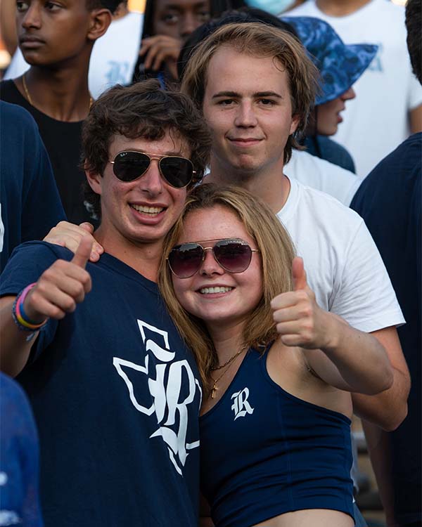 Three people wearing Rice shirts smile and pose with a thumbs up in the stands of the football field.