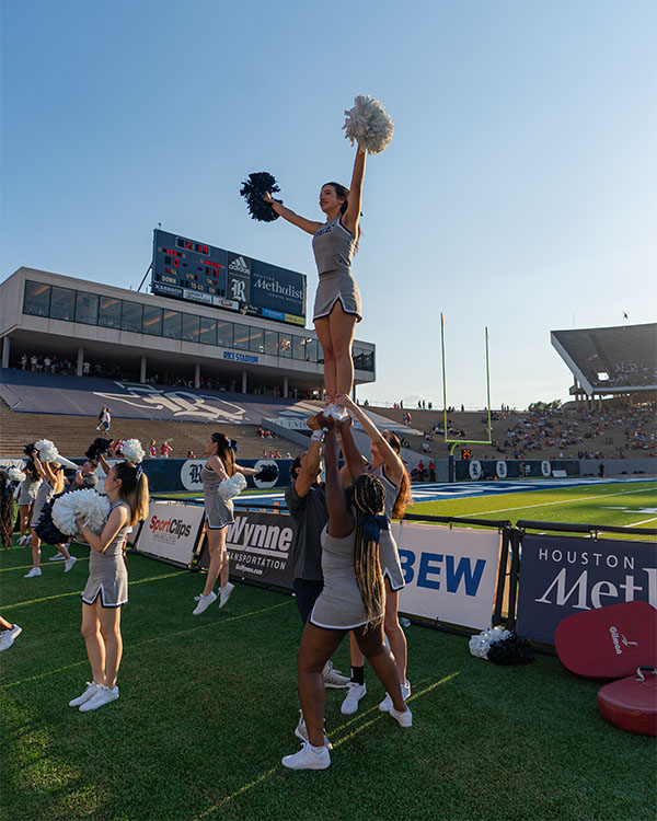 Three cheerleaders hold up a fourth with her arms outstretched in a v, waving her blue and white pom poms.
