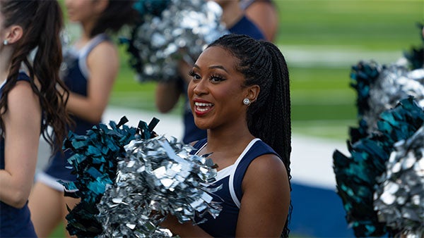 A Rice member of the dance team smiling and holding silver and blue pom poms is standing in the center of the picture.