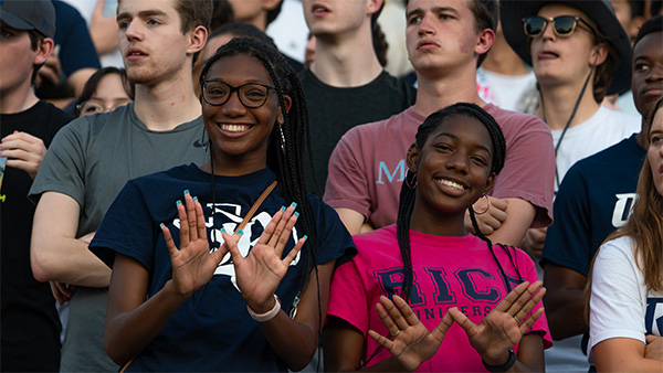 Two students in the stands wearing Rice shirts hold up the Owl Hand Sign with their thumbs crossed and palms facing out