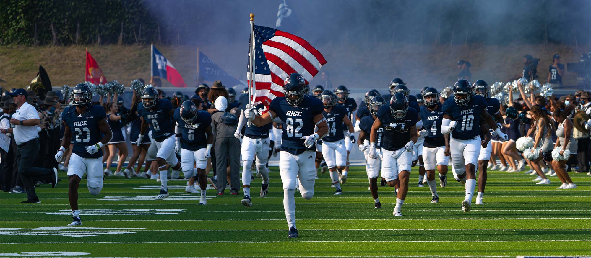A group of football players run towards the camera on the football field, with the one in front carrying an American flag.