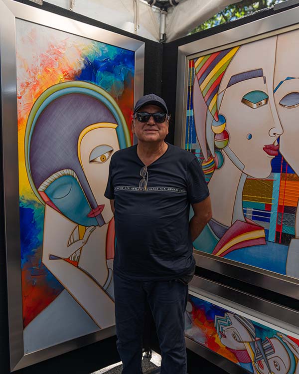 The artist, Julio Garcia, stands in front of his paintings, which are abstract portraits painted with primary colors.