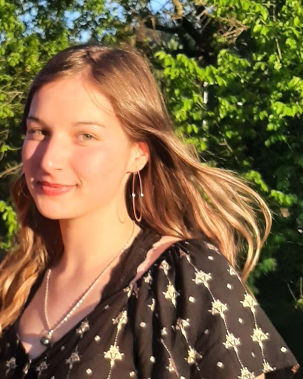 Grace poses outside for a headshot with a soft smile in front of a tree with bright green leaves.