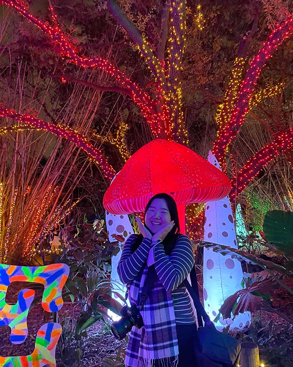 Sarah poses in front of a lit up mushroom and some lit up Christmas trees