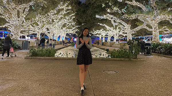 Grace poses in front of a long walkway lined with trees that are covered in white Christmas lights.
