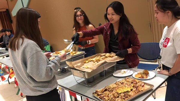 One female student wearing gloves serves breakfast food to another female student holding out her plate.