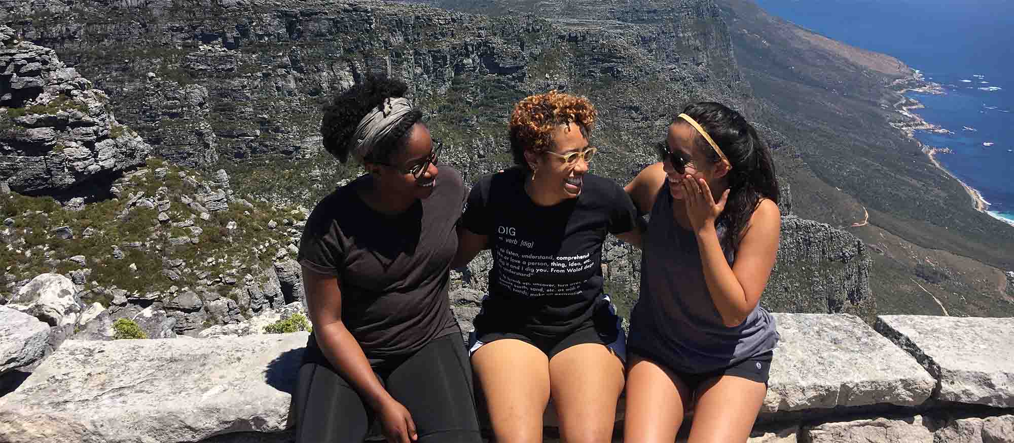 Three female students sit on a wall facing each other with a mountain range behind them.