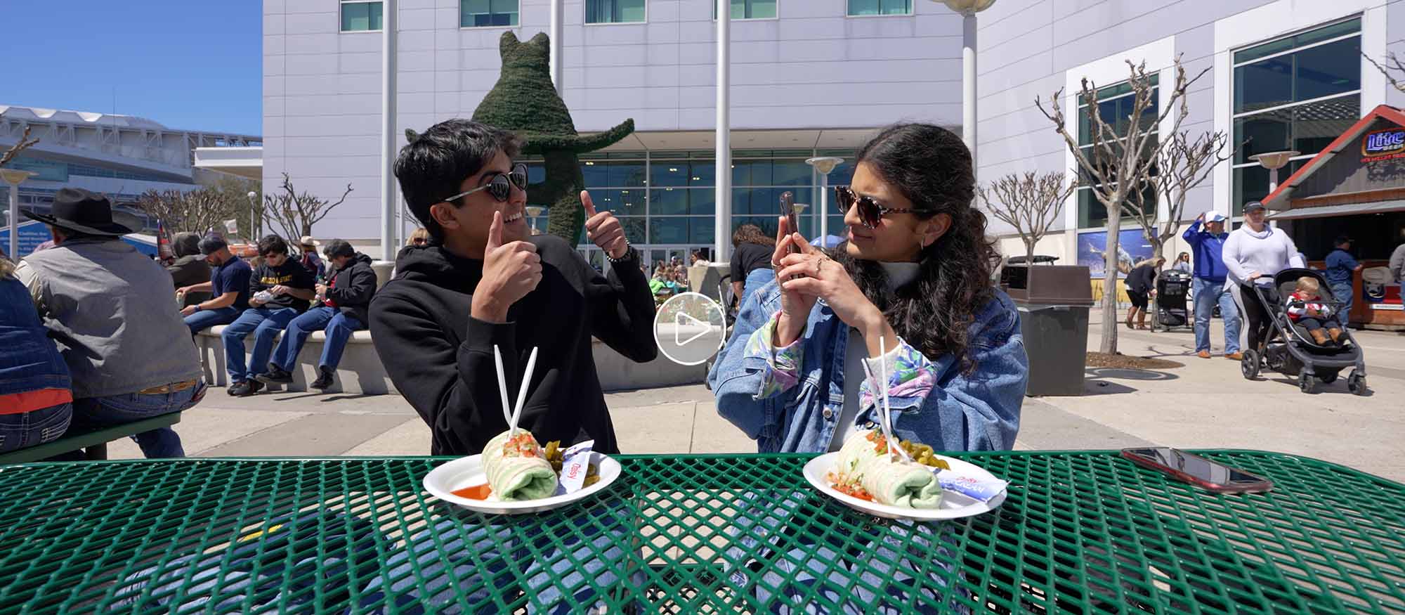 Naman, sitting at a picnic table on the left, turns smiling and holding two thumbs up to Devika on the right, who is taking a picture of him with her phone. There are two large burritos on plates in front of them.