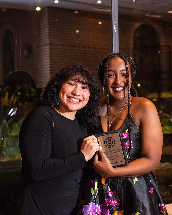 A Black student at Rice University smiles with an administrator with a plaque commemorating an award she's just won at the Black Excellence Gala.