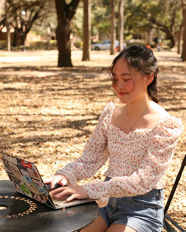 A picture of Emily M., an intern for the Houston Asian American Archive, sitting at a table outdoors working on her open laptop.
