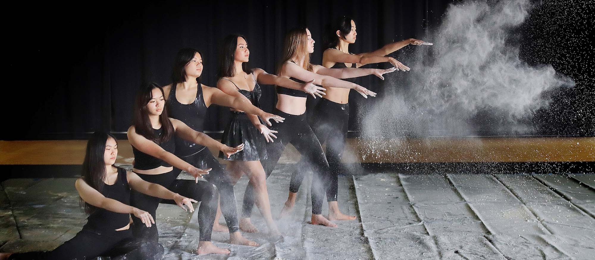 An all female group of six contemporary dancers pose in a line with their hands out, after throwing chalk which makes a cloud to the right of them.