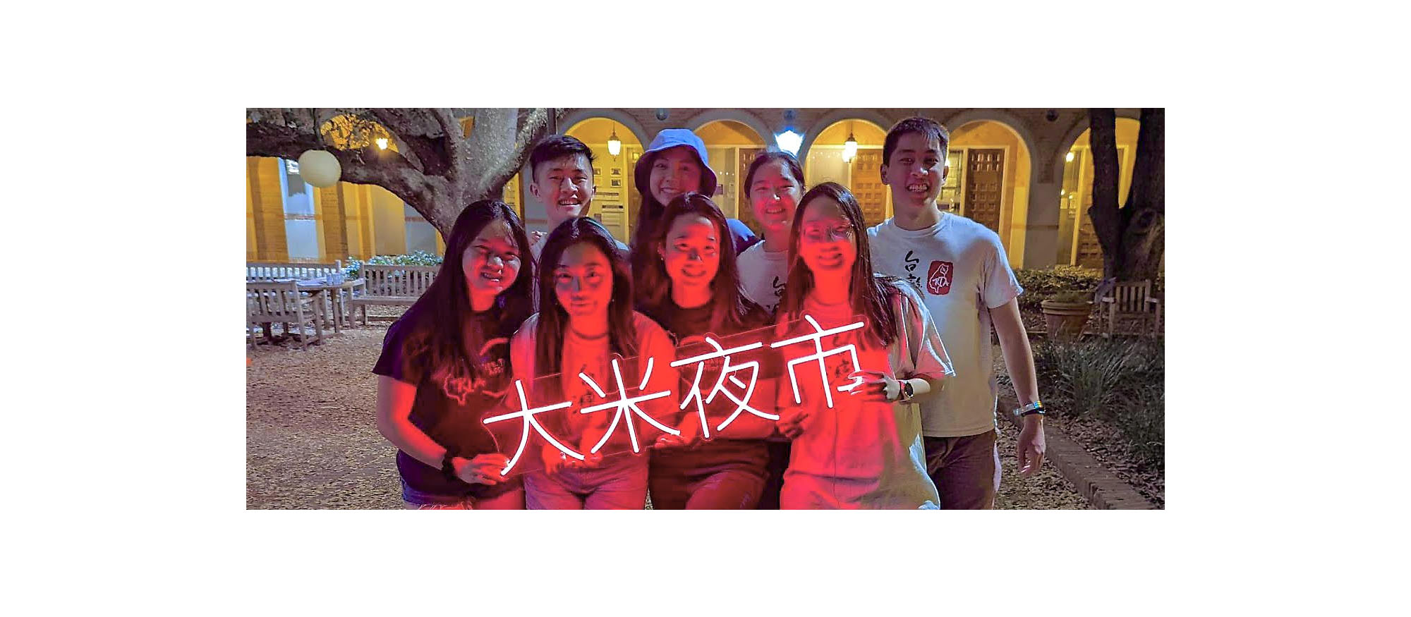 A group of male and female students from the Rice Taiwanese Association smile and pose with a neon sign that says "Rice night market" in Mandarin.