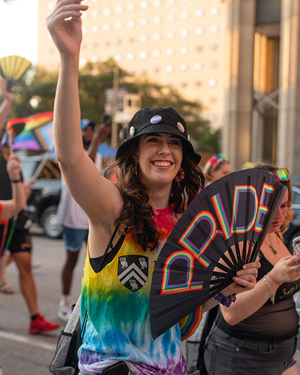 A member of the Rice community walking in the parade waves at the parade watchers and holds up a fan that says "Pride."
