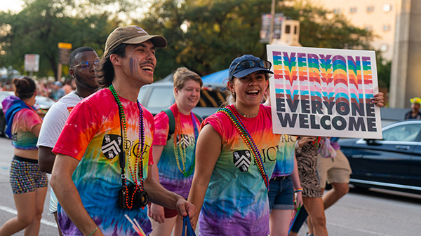 Two students wearing Rice Pride shirts walk along the parade route holding a sign that says Everyone Welcome in the pride flag colors.