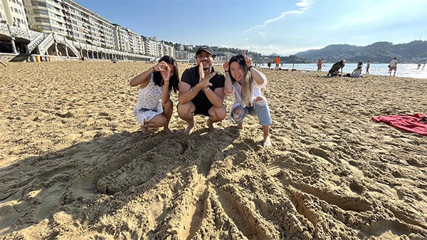 Linda and two other students are crouching on the beach next to the letters "BKR" spelled out in the sand, which stands for Baker.