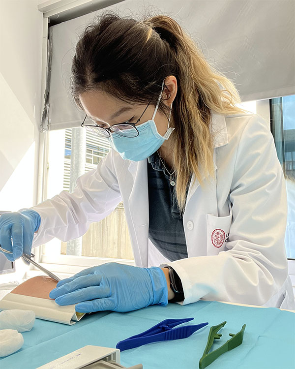 Linda is shown in the picture wearing a surgical mask, a white lab coat and blue gloves as she learns how to suture.