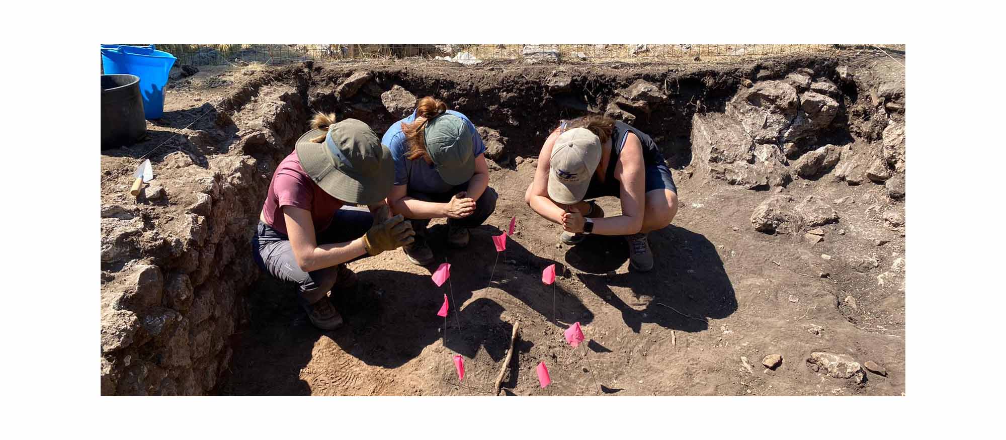 A group of students are squatting in the trench while holding out their hands to pink flags on the ground arranged in a circle.