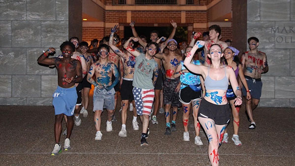 A group of students from Martel walks through the Martel sallyport covered in paint and with their arms raised, ready for an O-Week event.
