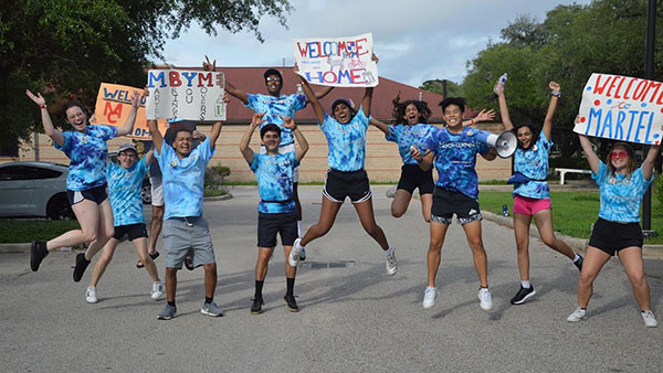 A group of students wearing matching blue tie-dyed O-Week T-shirts jumps up in the air holding signs above their heads that say things like "Welcome to Martel College."