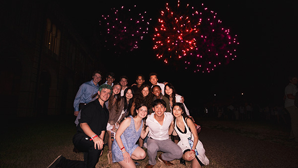 A group of students smiles for a photo at nighttime with a red firework in the background.