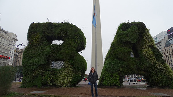 Brendan stands in front of two large shrub designs of the letters B and A for Buenos Aires as well as the Argentinian flag flying above.