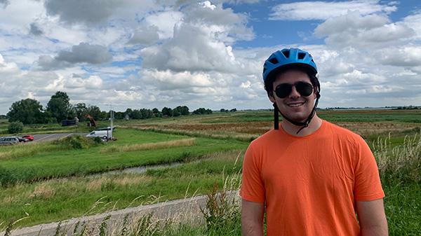 A picture of Zach wearing a bike helmet and posing in front of a large, green, field of grass and with a blue sky overhead.