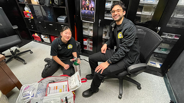 One student sits in an office chair to observe another student who is sitting on the floor with a fire extinguisher and an open bag filled with equipment.