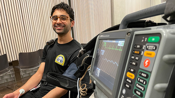 In the background of the photo, Anthony sits with a wire stuck on his arm that is attached to a monitor, pictured in the foreground, taking his vitals.