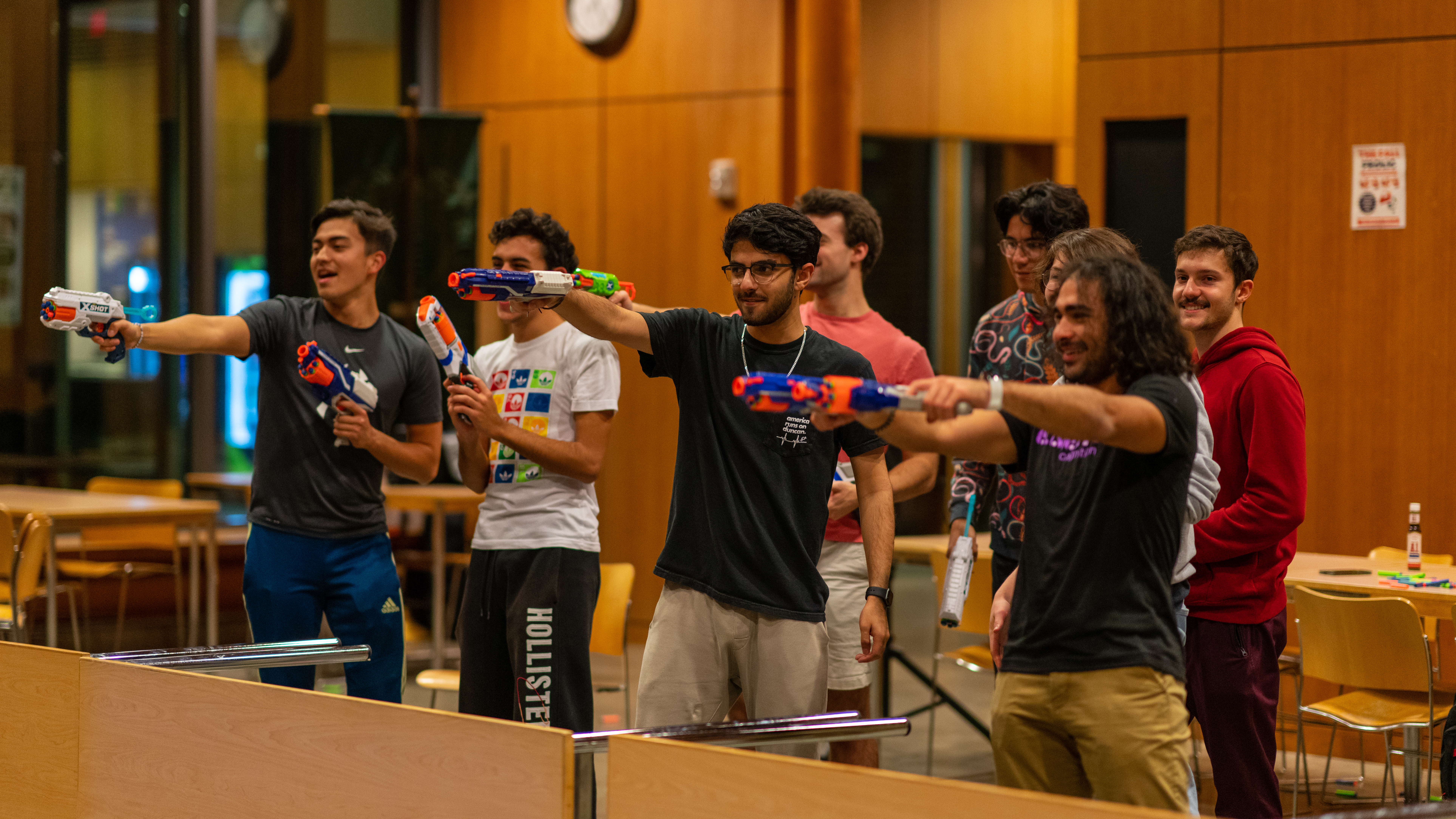 A group of students standing behind a table flipped to it's side, pointing their nerf weapons across the room.