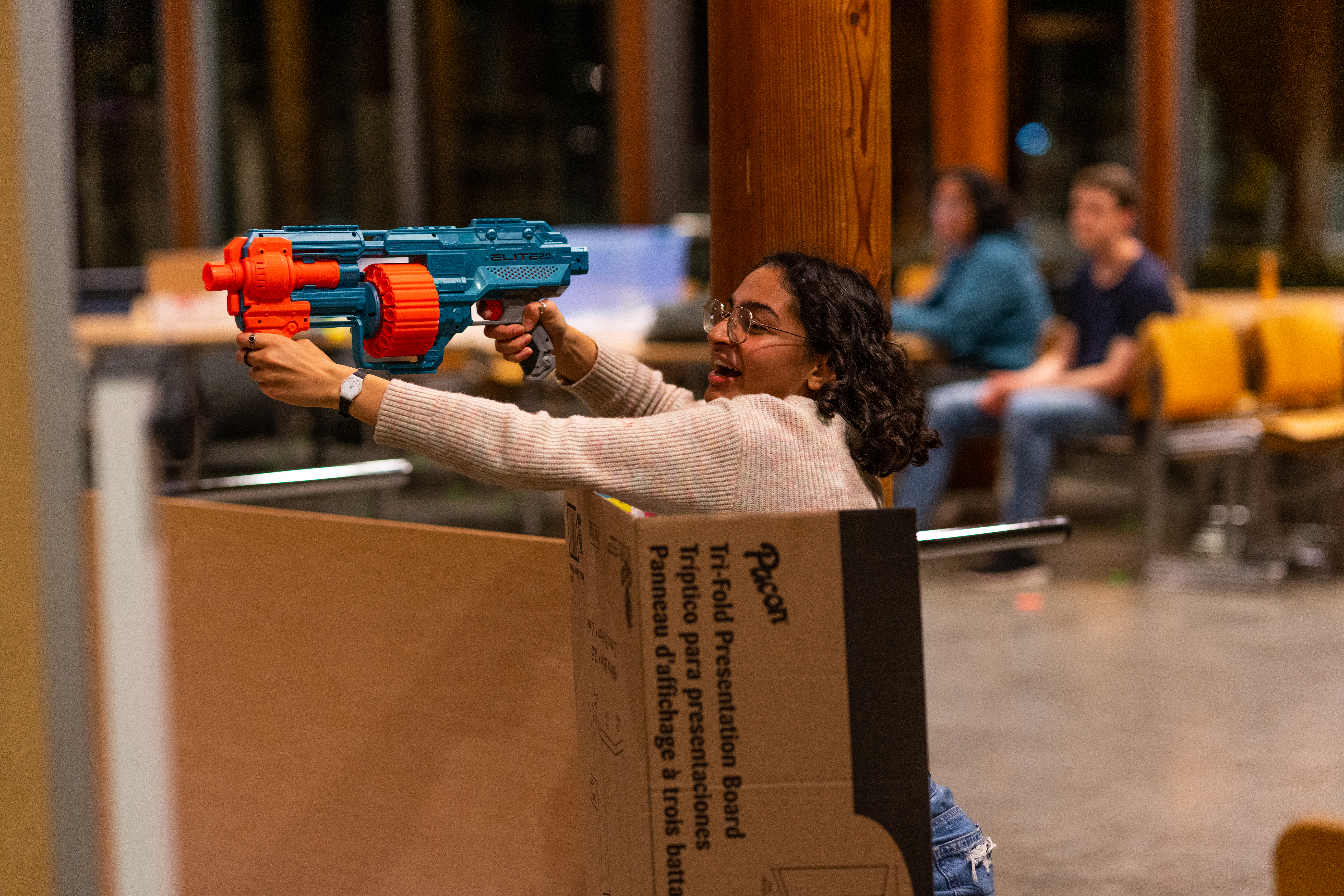 A female student holding a nerf weapon, reaching over the top of a cardboard presentation trifold and aiming across the room.