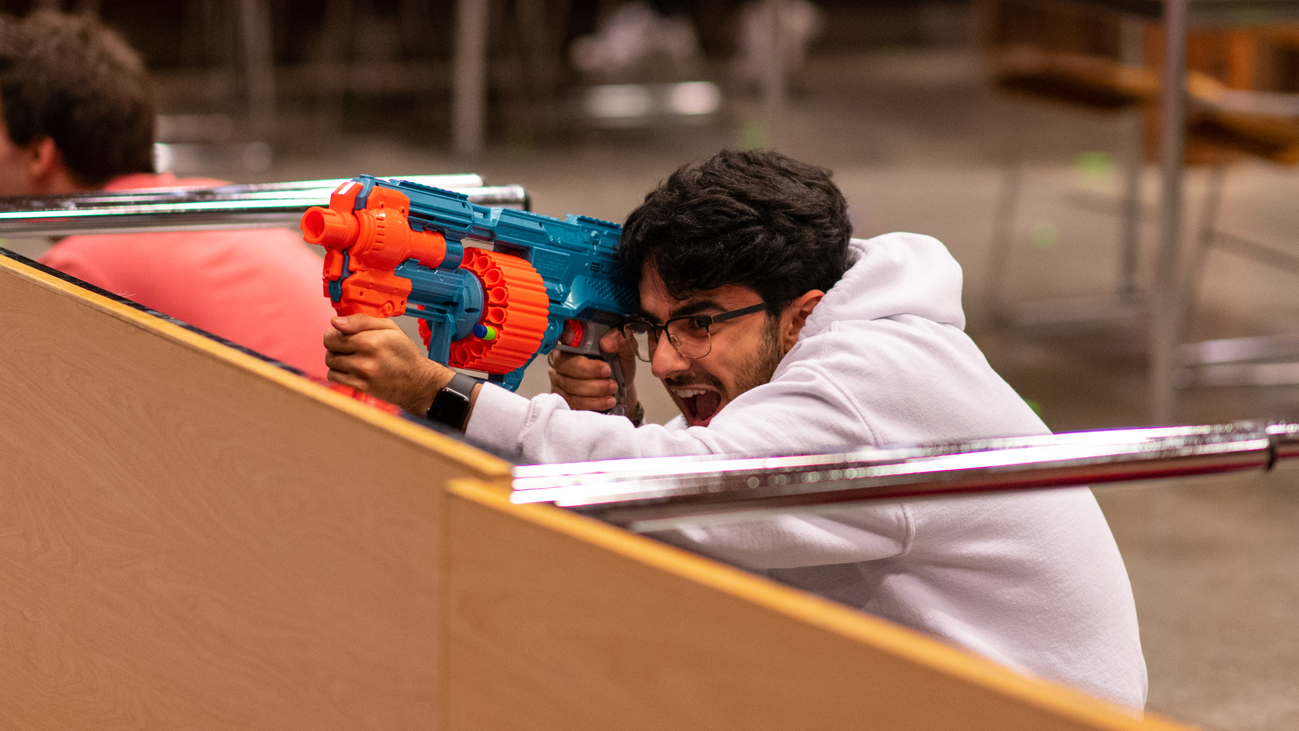 A male student holding an orange and turquoise nerf weapon near the left side of his face while crouching behind a table that has been flipped to its side.