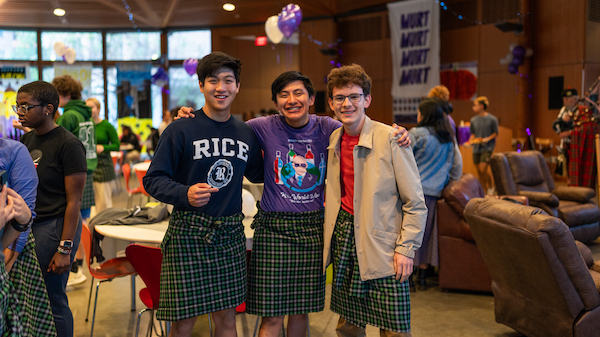 Three students wearing kilts and standing next to each other in a single row while smiling.